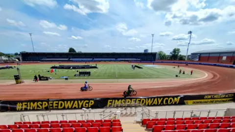 BBC / Giulia Bould A race track with red seating in front. Motorcycles can be seen on the red, dirt, track. There is a circular grass patch in the middle of the track. It is a sunny day with blue skies.