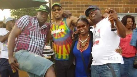 Three men and a woman in brightly coloured clothing hugging and smiling, one with a beer bottle in his hand.  