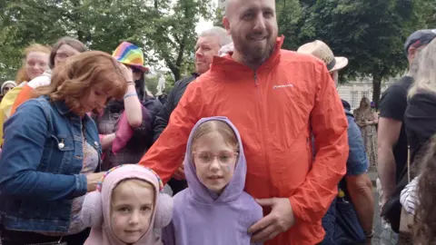 Man with his arms around two young girls amongst a crowd