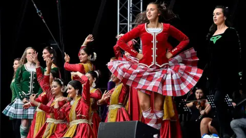 A large stage with a black background has a number of women taking part in Irish dance. The women are wearing black, red and green outfits with matching checked skirts.