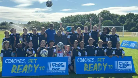 A group picture of smiling Rachel Yankey with the girl's team that she visited. Some of the girls are looking at footballs thrown in the air. It is sunny.