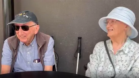 BBC Alan and Jean Robinson in sun hats sitting at a table near the new bridge