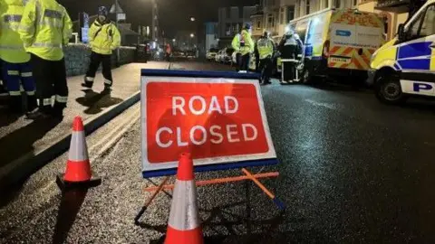 Road closed sign in Exmouth. It's dark with lots of emergency workers walking around.