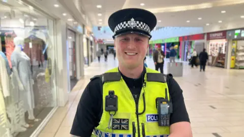 Mark Ansell/BBC PC Jake Hale smiling at the camera. He is in Frenchgate Shopping Centre in Doncaster and is wearing a police hat and high visability police vest which holds his walkie talkie. He has tatoos on his right arm.