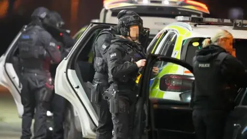 Four armed police officers standing behind police vehicles. There is also a woman with a police vest on in the foreground.