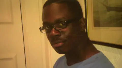 Family handout Dwayne Simpson, a young man, with short black hair, black rimmed specs and a plain grey t-shirt, looks at the camera. He is in a living room with a painting behind him and a white door to the left.