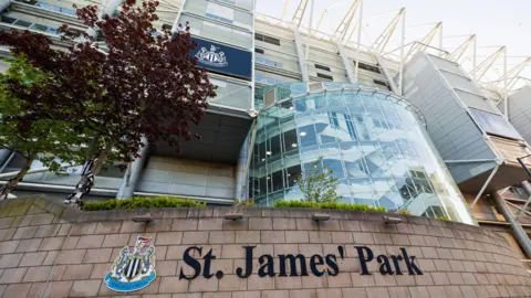 Supplied St James' Park sign on a brick wall on the Newcastle United football stadium. There are large curved glass windows.