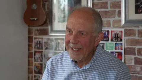 Shaun Whitmore/BBC Steven, a man who is wearing a white and blue chequered shirt. He is looking away from the camera and has pictures and a guitar on the wall behind him. 