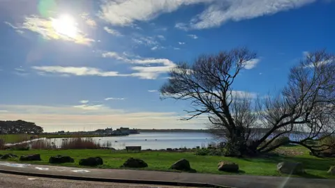 MarkieB The sun breaks through a cloud in an otherwise blue sky overlooking water and greenery. 
