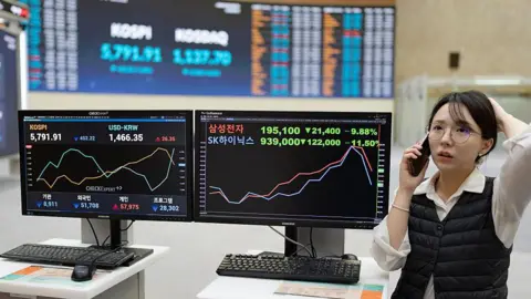 A woman in a black puffer vest scratches her head while speaking over the phone. She is standing in front of an electronic scoreboard at the Korea Exchange in Seoul, South Korea. The screens display sharply fallen closing indices.