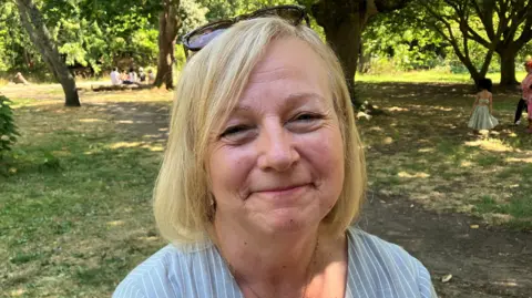 Melani Smith is photographed outside in a park. Grass and trees can be seen behind her. She has short shoulder length blonde hair and is wearing sunglasses on top of her head. She is smiling and looking at the camera.
