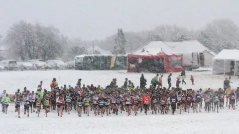 A crowd of people running in the distance on a field covered in snow. Snow is coming down heavily around them. The cars parked in the distance are completely covered. There are tents and portaloos at the back of the field.