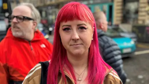 Courtney MacKinnon A woman with pink hair stands in the middle of the street with a light brown Adidas coat on