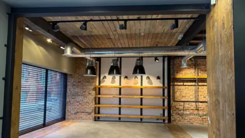Sarah Farrell-Foster Inside a ground floor building. To the right is blue shutters down and at the back there is a exposed brick wall with wooden beams and black pendant lighting hanging down from the ceiling. 