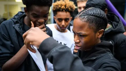 Getty Images A student, who is wearing a black jacket and has a braid in his hair, looks quizzically at his GCSE results slip. His friends look on, one of them holding his hand to his mouth.