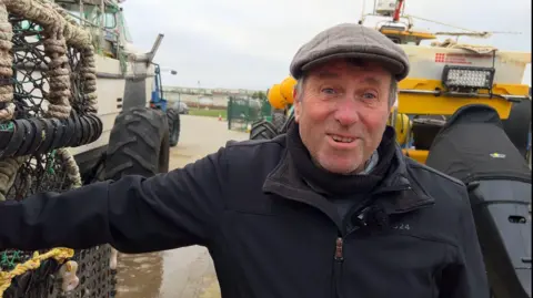 Alan Stead is wearing a black coat and and a grey cap, he is leaning against lobster pots and two vessels are visible behind him.