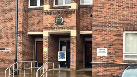 Qays Najm/BBC The entrance to Norwich Crown Court. It has a red brick facade with stonework and the royal crest, and the words "The Law Courts: Crown and County Court" above the door. A notice is on the wall and another notice is on a stand at the entrance.