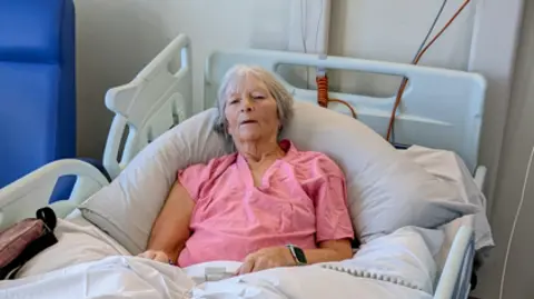 A woman in a pink top with short grey hair lies in a hospital bed.