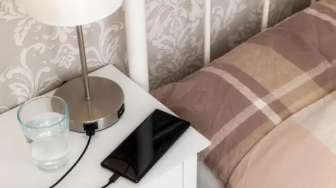 Getty Images/Thomas Faull A smart phone charging on a white bedside table.  There is a silver table lamp with a plain white shade next to the phone.  A glass tumbler half filled with water is also resting on the table.  A brown and beige checked duvet set covers the bed which has a white metal frame.