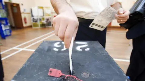 A hand places a ballot paper into a black ballot box at a polling station. The box has a numbered label on its lid and a red security seal attached to the slot, with a voter holding paperwork standing behind it in a hall.