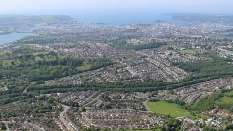 An aerial view of a large cityscape with green areas and sea beyond.