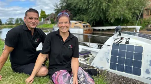 Ian and Jooles Paillin-Dean sat on the grass by the water next to their boat