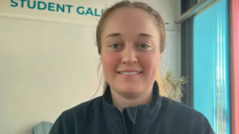 Poppy Ludgate is in an office room, wearing a dark blue fleece, she is smiling at the camera.