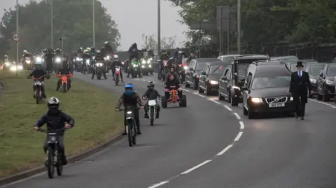 Dozens of bikers and youngsters on scrambler bikes ride alongside the funeral cortege. The bikes and cars have their lights on in the gloom. 