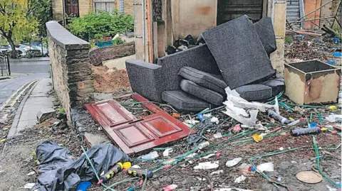 LDRS A run-down outdoor area beside a brick building that has been filled with dumped furniture and rubbish. Several large sofa cushions and upholstered sections are piled against a damaged doorway, while a red wooden door lies flat on the ground nearby. The ground is covered with scattered waste, including plastic bottles, cables, and broken materials.