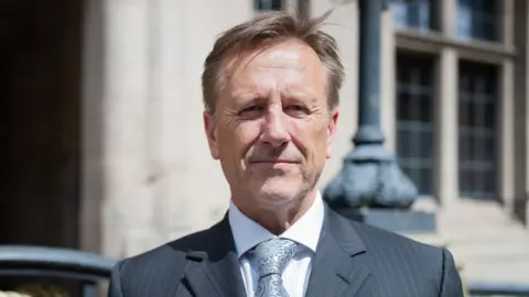 Martin stands infront of the county council building in Stafford on a sunny day, smiling at the camera. He is wearing a black suit and grey tie. 