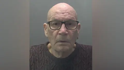 Cambridgeshire Police Custody photograph of David Newton, who is clean shaven and bald, looking into the camera, wearing dark glasses, a ring-shaped earing and purple jumper