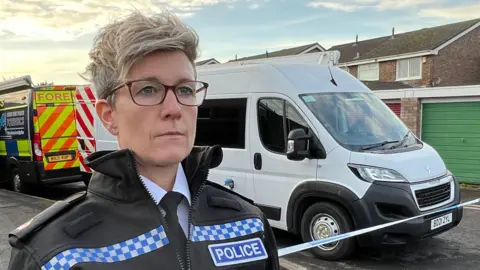 A police officer with short blonde hair and glasses in her uniform of black with blue and white checks on the shoulder lapels. She is standing on a residential street in front of a police cordon.