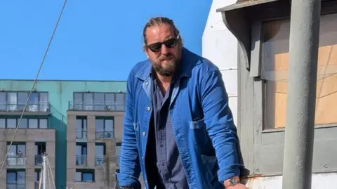 Julien Jourdain A man stands on a boat on a sunny day. He has long hair that is tied back and he is wearing a blue denim jacket with a darker blue shirt underneath. He is also wearing sunglasses.