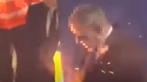 Instagram A still image showing sparks and smoke as a flare strikes a steward, in a suit and tie, standing against a blue wall.