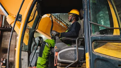 A man in work clothes, including a yellow hard hat, sitting at the controls in the cab of a digger.