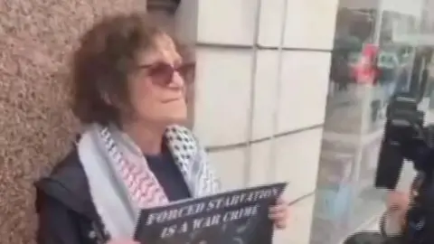 Screengrab of dark haired woman holding a sign that says "forced starvation is a war crime". She is wearing sunglasses and a black and white scarf, probably a keffiyeh, and has a dark coat on. 
