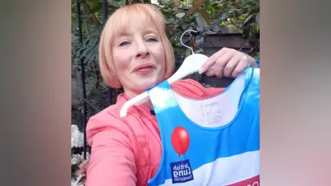 Chloe Slasberg, wearing a pink jacket, holding up a blue and white running top on a white hangar.