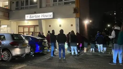 A group of people wearing winter clothing stand outside a block of flats in the dark. There is a sign on the building which says 'Barton House'. 