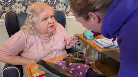 The Birches A woman with blonde hair and a blue flower in her hair sits in a chair. Another woman wearing a purple jumper is standing in front of her holding a tortoise. The two women are having a discussion.