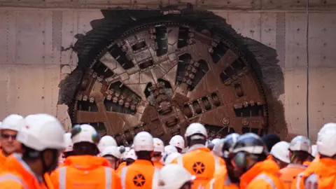 PA Media A huge metal circle is protruding out of a piece of land. The metal is bronze in colour. Dozens of people in white hard hats and orange high-vis jackets are standing in front of it
