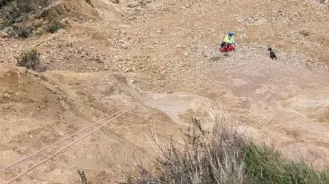 Lyme Regis Coastguard A view from above of a climber in hi-vis gear and attached to ropes near a dog on a large stony plateau
