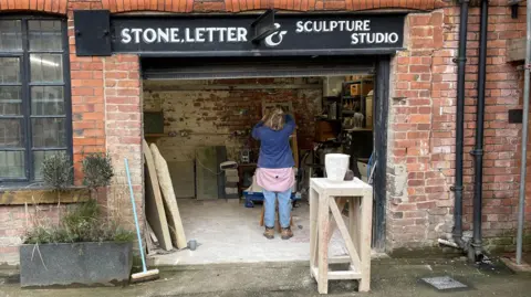 Mark Ansell / BBC An artist's studio viewed from the outside. A red-brick building with a black steel sign and a wide door way from which sculptor Lily Marsh can be seen working from behind.