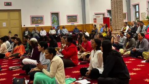 A group of men and women sit in a large room on red carpet. Their knees are crossed and they are holding their hands in prayer. 