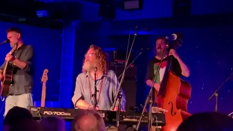 Three members of a band, one on guitar, another on keyboards and another on bass perform on stage.