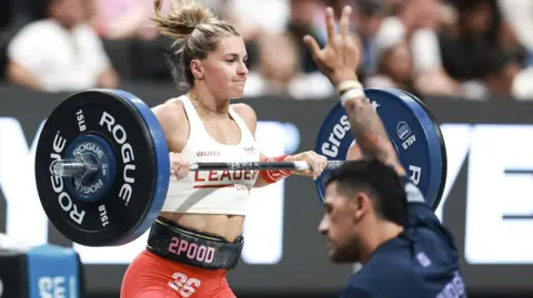 CROSSFIT GAMES Cringle lifting a large weight. She is wearing a white vest with leader in red on the front.