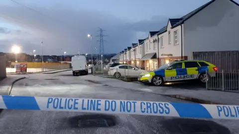 Police tape is in the foreground of the image, behind the tape sits a parked police car, which is in front of a row of houses. A white van and a number of parked car are on the road. the road is icy.