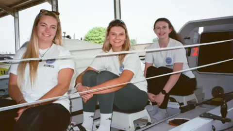 Liv Stocks pictured with Jenna Vincent and Meg Neely. They are in a boat and are wearing white matching t-shirts with a logo. They are all smiling at the camera.