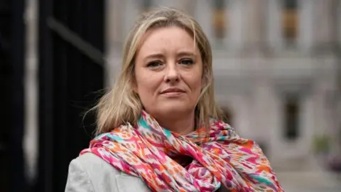 PA Media Cahill has long blonde hair, she is looking directly at the camera. She is wearing a brightly patterned pink, orange and blue scarf..