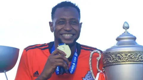 Vipers Sports Club/X Abubabakar Lawal, in a red football jersey, holding a medal and a cup by his side