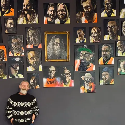 Simon Gee Simon Gee, with a grey beard, wears a black and white striped jumper and is standing in front of a dark wall where about 20 of his portraits are hung. Many of them depict people wearing bright yellow and orange hi-vis jackets. 